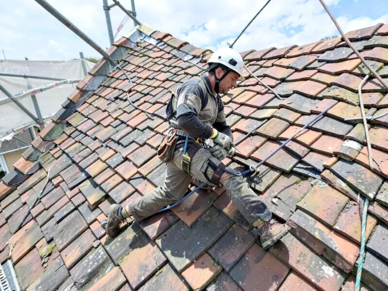 Dachdecker verlegt Dachziegel auf einem Steildach, angeseilt und mit Helm